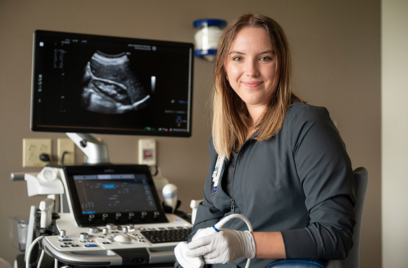 Photo of NDSU sonography student Lydia Fortin standing next to sonography equipment in a patient care area, holding a transducer, wearing gloves preparing for a sonography exam.